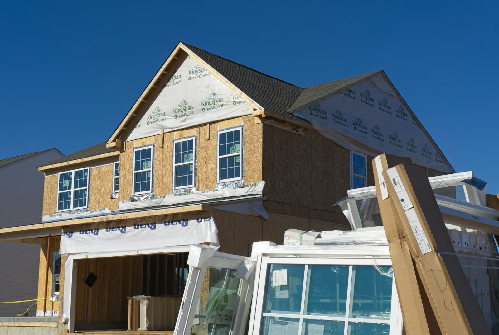 A single-family home under construction — sheathed and wrapped, with new windows stacked out front ready to install.
